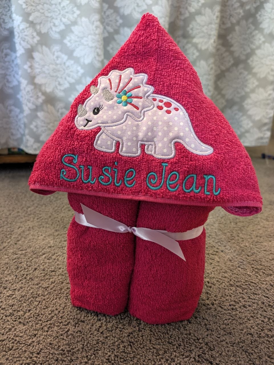 Hot pink hooded towel folded and tied with a ribbon, a polka-dot triceratops appliqué and an embroidered name on the hood