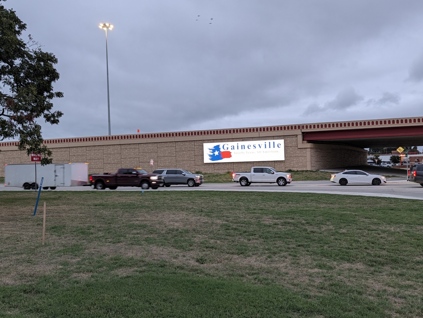 The Gainesville welcome sign on the I-35 overpass, seen across the grass with traffic on the frontage road.