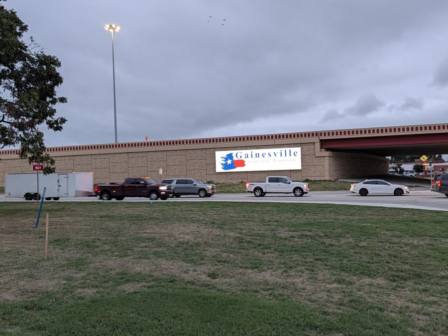 The Gainesville welcome sign on the I-35 overpass, seen across the grass with traffic on the frontage road.